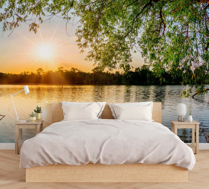 A surreal bedroom setting overlooking a calm lake with a bright sunrise, featuring Lakeside Branches Wallpaper Mural by Decor2Go Wallpaper Mural on the bed with white linen and side tables with minimal decor under a tree canopy.