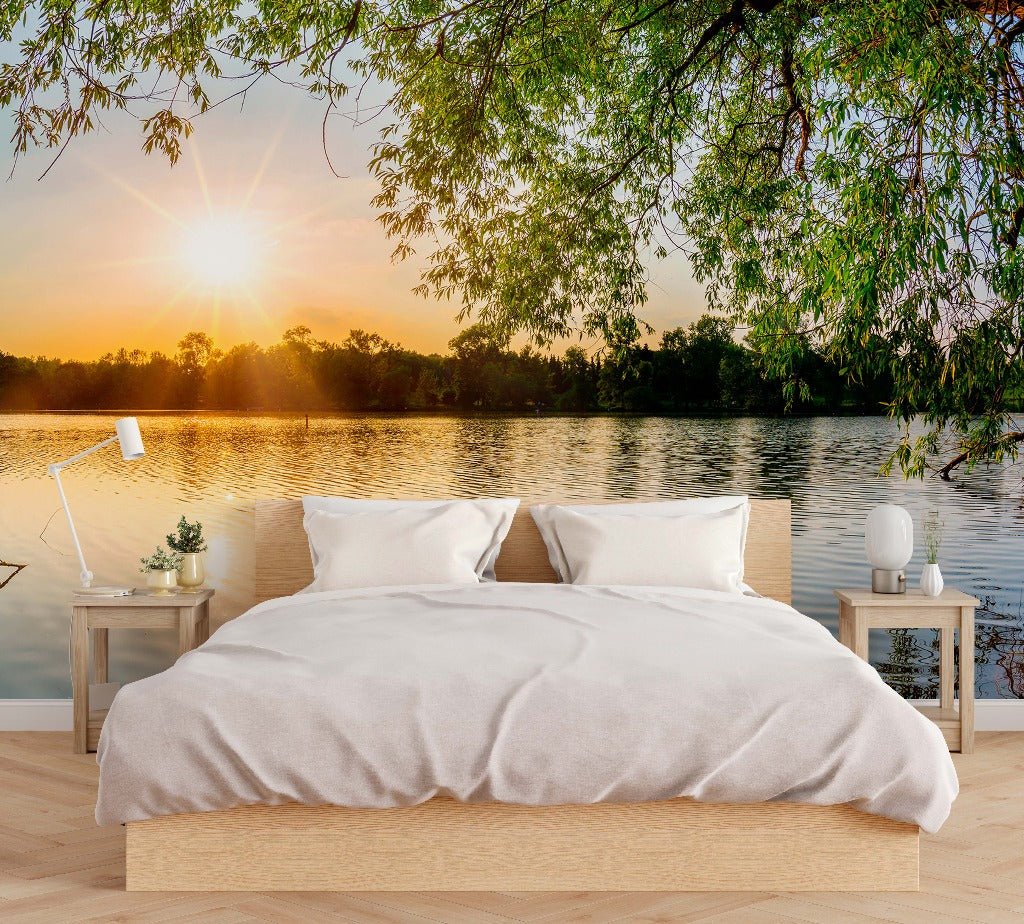A surreal bedroom setting overlooking a calm lake with a bright sunrise, featuring Lakeside Branches Wallpaper Mural by Decor2Go Wallpaper Mural on the bed with white linen and side tables with minimal decor under a tree canopy.