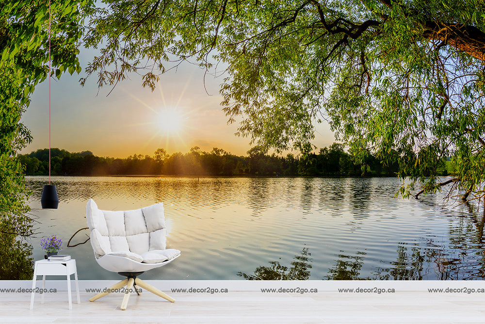An outdoor scene blending nature with home decor, featuring a stylish white chair and a small table set by a calm lake surrounded by lush green trees under a brilliant sunset, featuring the Lakeside Branches Wallpaper Mural from Decor2Go Wallpaper Mural.