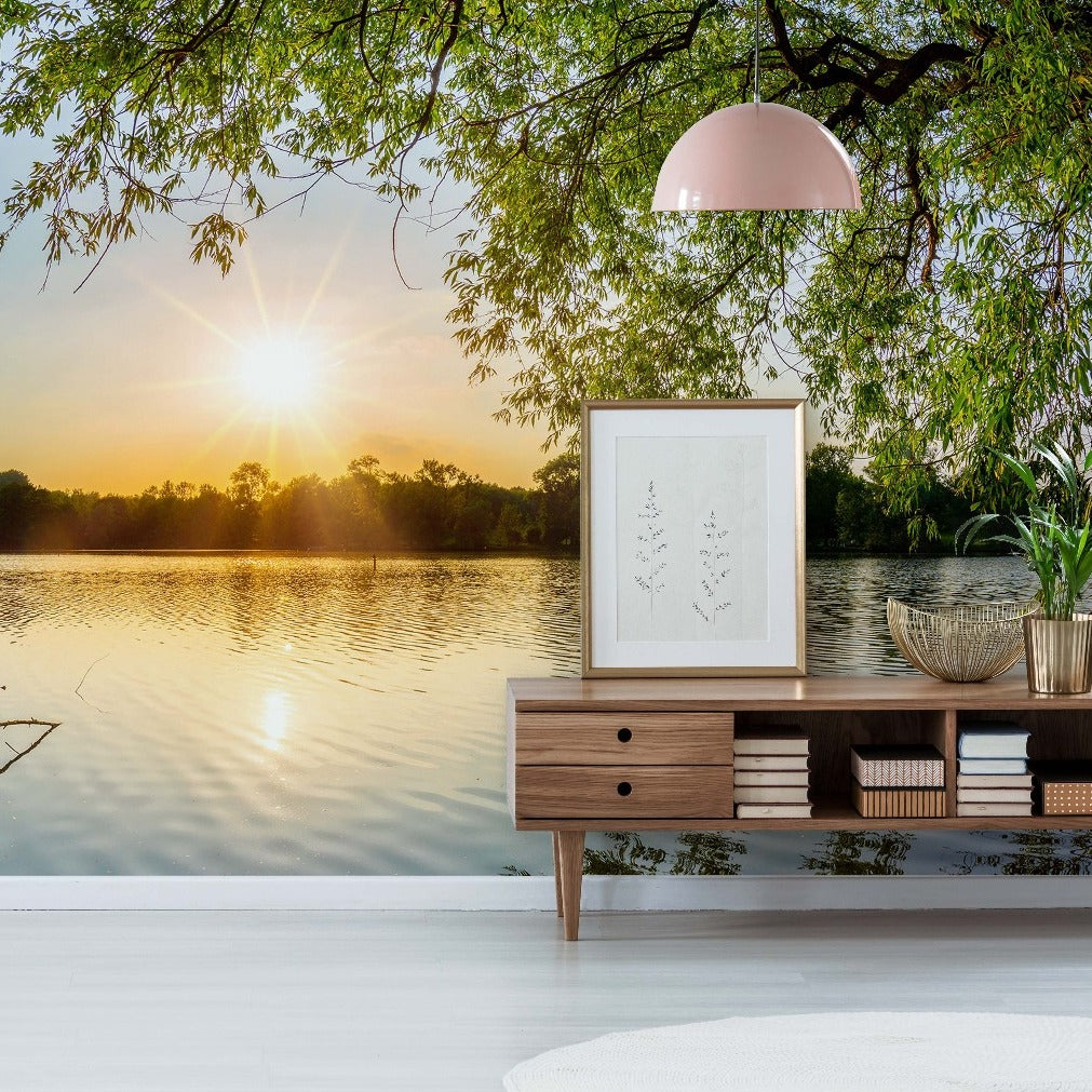A serene lakeside view at sunrise seen from a stylish room with a wooden sideboard, decorative items, and a framed Lakeside Branches Wallpaper Mural from Decor2Go Wallpaper Mural, all under a soft pink hanging lamp.