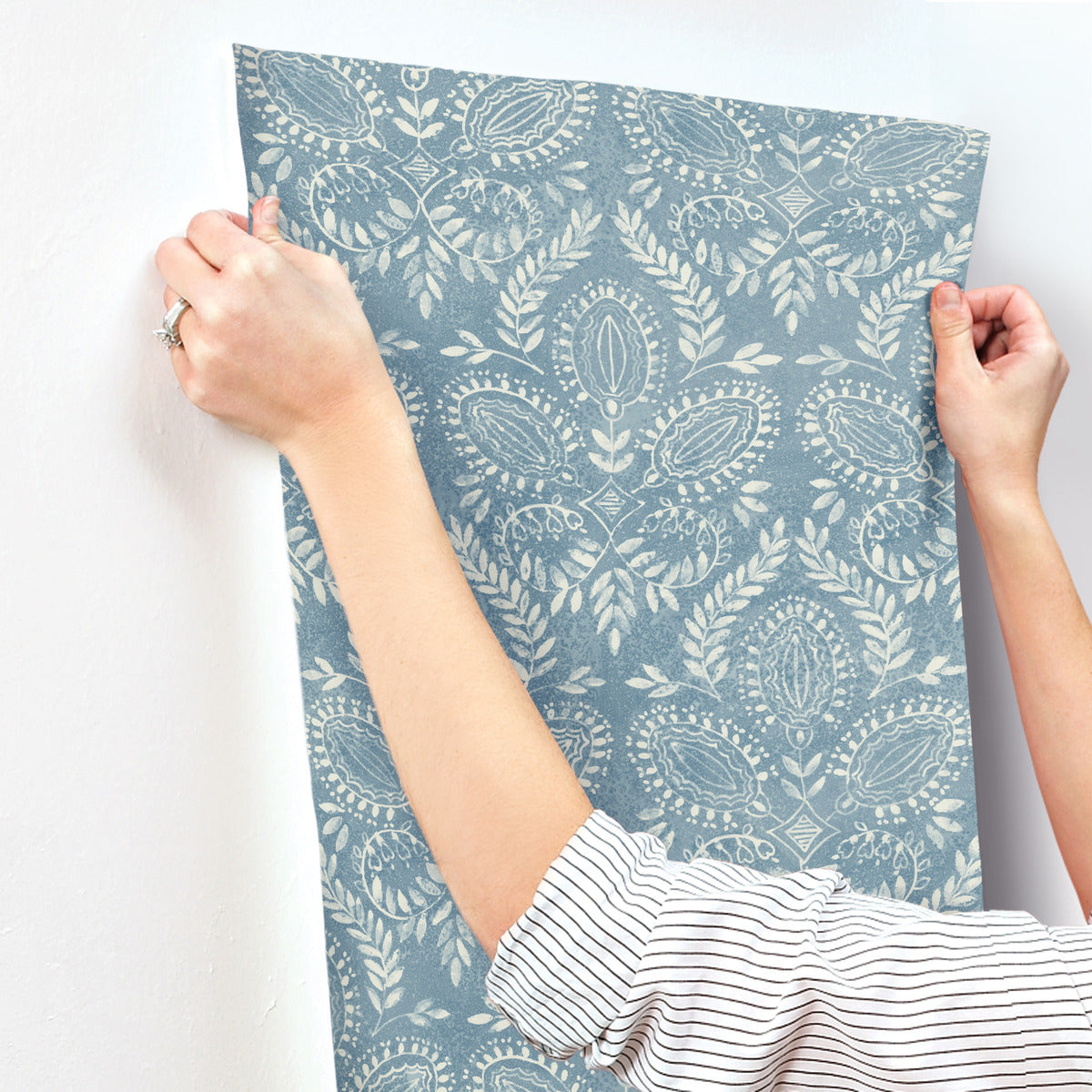 A person wearing a striped shirt is holding a piece of York Wallcoverings' Laurel Damask Wallpaper Blue (60 SqFt) against a white wall. This nature-inspired design features an intricate floral pattern with leafy details, capturing the essence of the wallpaper's light-colored aesthetic.