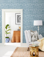 A cozy living room showcases the York Wallcoverings' Laurel Damask Wallpaper in blue patterns. A white armchair with decorative pillows and a fluffy throw is positioned next to a small gold table. Through the doorway, a potted plant enhances the nature-inspired design, while a wicker table and abstract art are visible in the background.