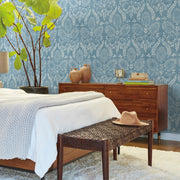 Bedroom - A stylish showcases a bed adorned with white and light gray blankets. A wicker bench holds a tan hat, while the wooden dresser is arranged with books and vases. The nature-inspired design is accentuated by a large leafy plant set against vibrant York Wallcoverings' Laurel Damask Wallpaper in blue