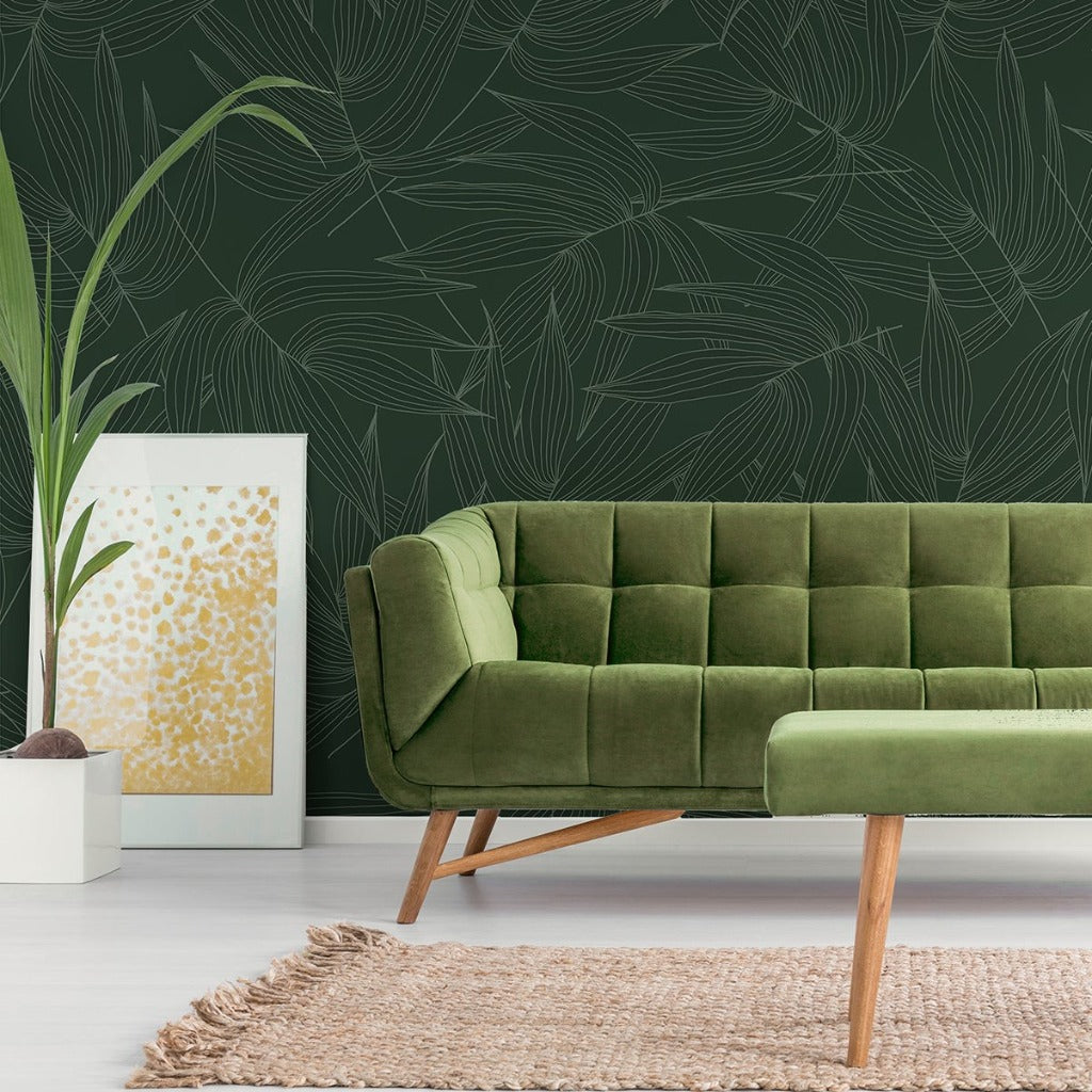 A modern living room featuring a plush green sofa against a dark green feature wall with a Leaves lines pattern Mural Wallpaper from Decor2Go Wallpaper Mural. A white frame with abstract art and a plant beside the couch, over a beige rug, complete the.