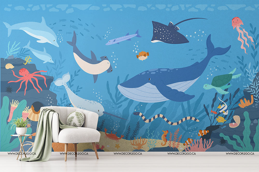 A colorful Decor2Go Life Underwater wallpaper mural on a room wall depicting a variety of sea creatures, including a whale, sharks, and jellyfish, accompanied by a cozy chair and stylish decorative elements.
