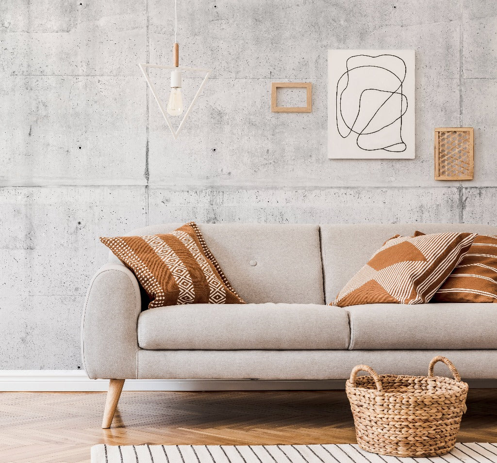 A stylish living room featuring a modern grey sofa with decorative brown pillows, set against a Decor2Go Wallpaper Mural with minimalist art and hanging geometric lights. A woven basket sits by the sofa.
