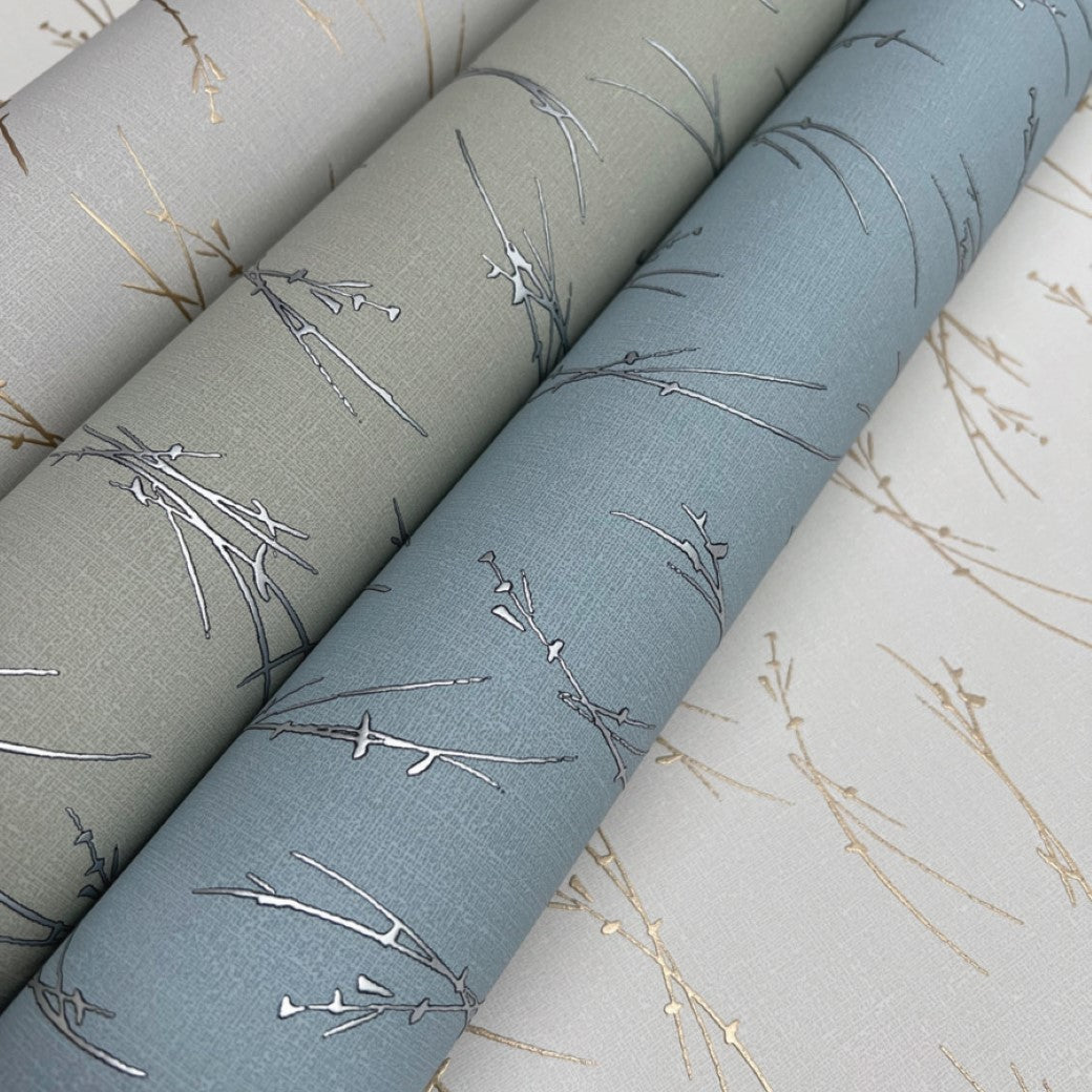 Three rolls of York Wallcoverings' Sprigs Wallpaper feature delicate branch patterns: one is a light tan with a metallic gold touch, another is sage green with silver accents, and the last is teal with silver branches, all offering elegant, soft grey backgrounds.