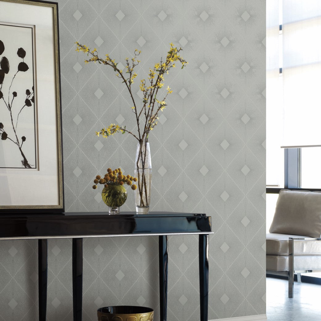 A modern interior features a black console table with a vase of yellow flowers and a bowl of decorative spheres. The room's design is enhanced by York Wallcoverings' Light Grey & Silver Harlowe Wallpaper, which has a metallic diamond pattern that complements the abstract artwork.