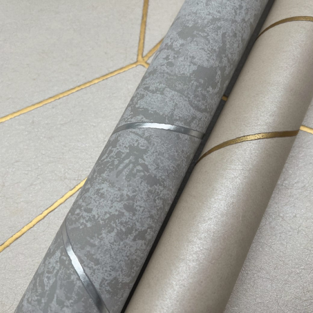 Two rolls from the York Wallcoverings modern metals collection are on a textured surface with gold geometric lines. The Light Grey & Silver Nazca Wallpaper features a metallic gray and silver pattern, while another roll is smoother beige with gold accents.