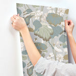 A person with rolled-up sleeves is applying York Wallcoverings Lotus Pond Cotton/Peacock Wallpaper Green, Pink (60 Sq.Ft.) featuring a botanical design with large leaves and flowers in shades of green, white, and beige on a light gray background to a white wall. The person's hands and wrists are visible, showcasing the easy installation and removal.