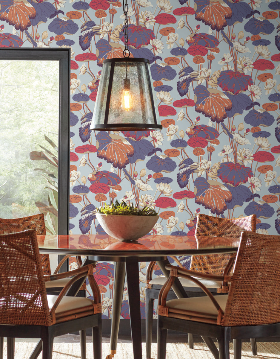 A dining area featuring a round wooden table surrounded by four rattan chairs. A metal pendant light hangs above the table, which holds a bowl with a plant. The wall behind is adorned with vibrant floral "Lotus Pond Cotton/Peacock Wallpaper Green, Pink (60 Sq.Ft.)" by York Wallcoverings in shades of blue, purple, and red.