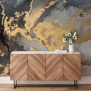 A modern wooden sideboard with a chevron pattern sits against a wall adorned with Decor2Go's "Luminous Marble" wallpaper mural. On top of the sideboard are a vase filled with white flowers, a candle, and a book titled "DESIGN." The contemporary space is enhanced by subtle metallic highlights that add to its artistic decor.