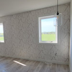 A bright room with light wood flooring and two windows features white walls and one adorned in York Wallcoverings' Gilded Marble Wallpaper. A geometric ceiling light and a single bulb enhance the room's sophisticated allure.