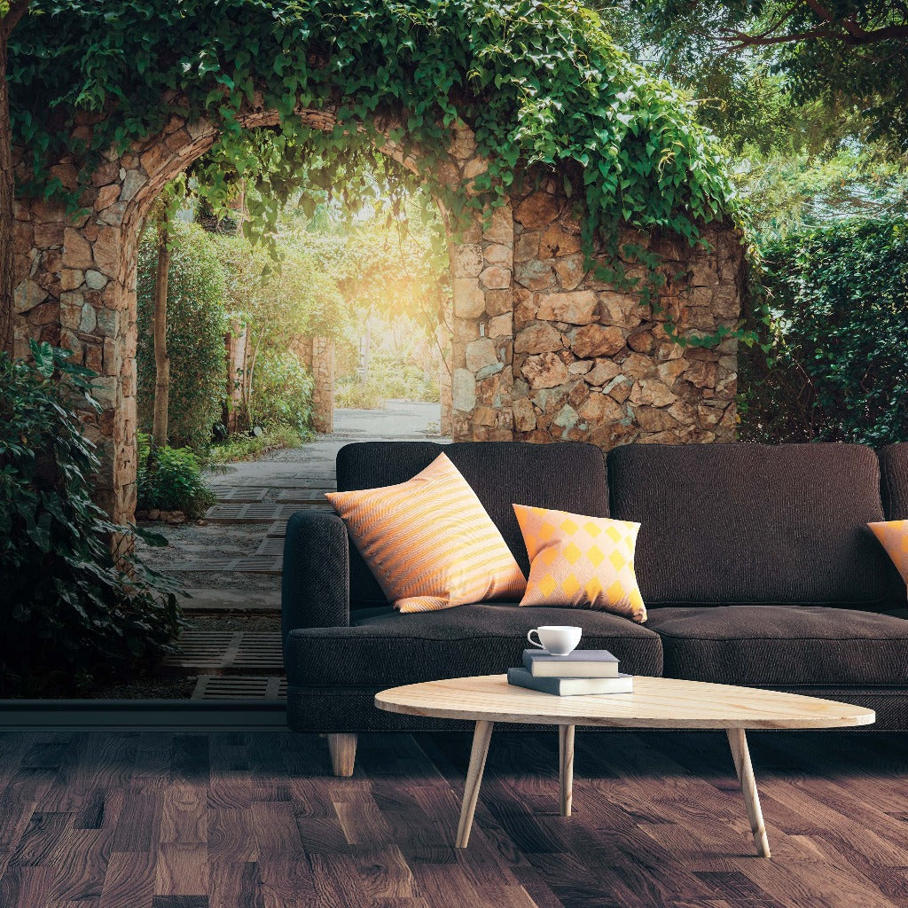 A modern black sofa with colorful pillows in a serene garden with a stone archway, lush trees, and a Decor2Go Wallpaper Mural. A coffee cup rests on a round table in front.