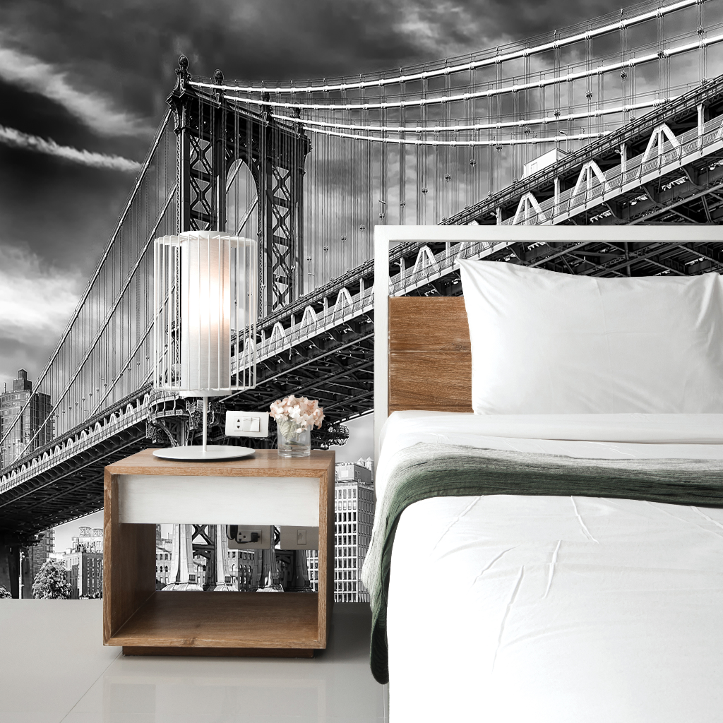 A bedroom with a large bed and a wooden nightstand featuring a lamp and flowers, with a dramatic Decor2Go Wallpaper Mural of the Manhattan Bridge on the wall behind it.