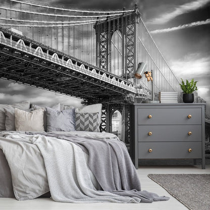 A bedroom with a surreal architectural design featuring a large Decor2Go Wallpaper Mural Manhattan Bridge Wallpaper Mural superimposed above the bed against a stormy sky backdrop, with modern furnishings including a dresser and plants.