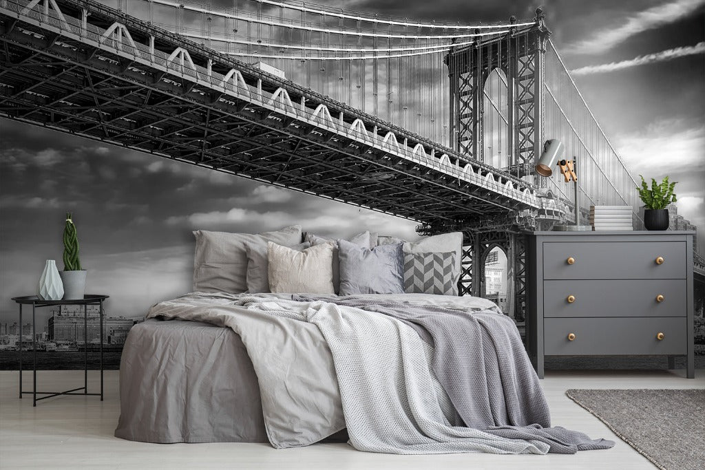 A bedroom with a surreal architectural design featuring a large Decor2Go Wallpaper Mural Manhattan Bridge Wallpaper Mural superimposed above the bed against a stormy sky backdrop, with modern furnishings including a dresser and plants.