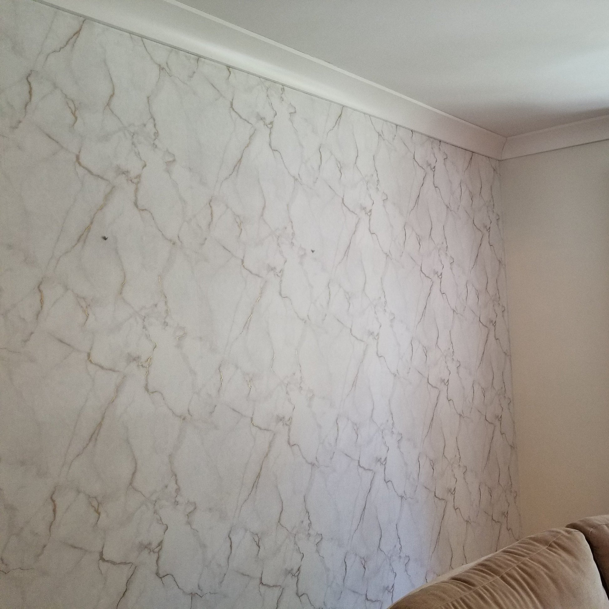 A living room corner showcases an elegant luxury beige sofa against York Wallcoverings' Gilded Marble Wallpaper. Non-woven curtains accent the window, where sunlight filters through, brightening the space alongside a floor lamp.