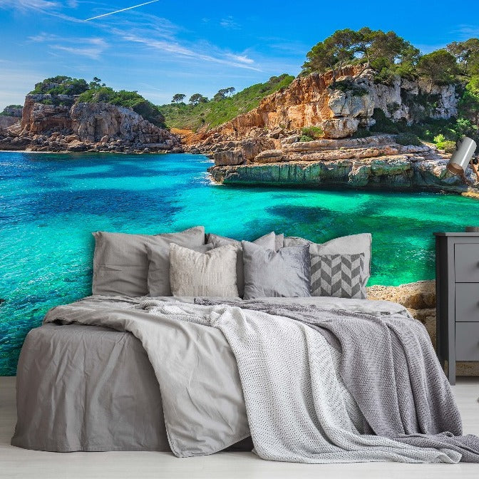 A bedroom setup with a bed and side tables in the foreground, facing a large window with a clear view of the stunning blue sea surrounded by lush green cliffs, enhanced by Decor2Go Wallpaper Mural.