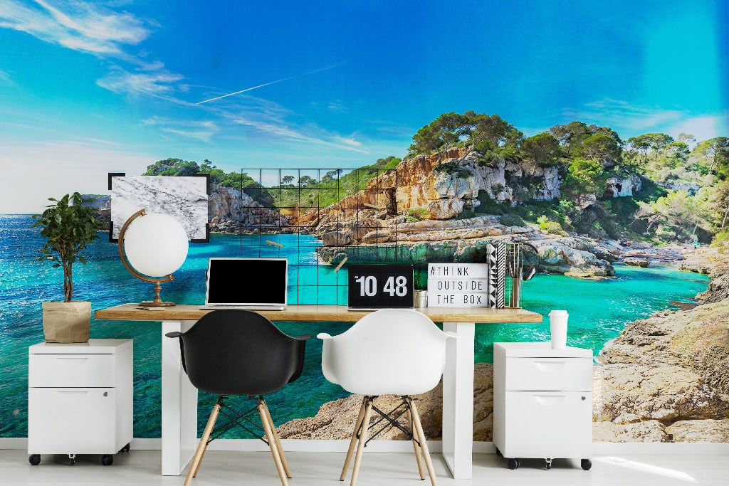 A modern home office setup with a large desk facing a wall-sized window overlooking a vibrant coastal scene with rocky cliffs and a turquoise sea. The desk features a computer, globe, and inspirational decor enhanced by Decor2Go Wallpaper Mural's Mediterranean Blues Wallpaper Mural.