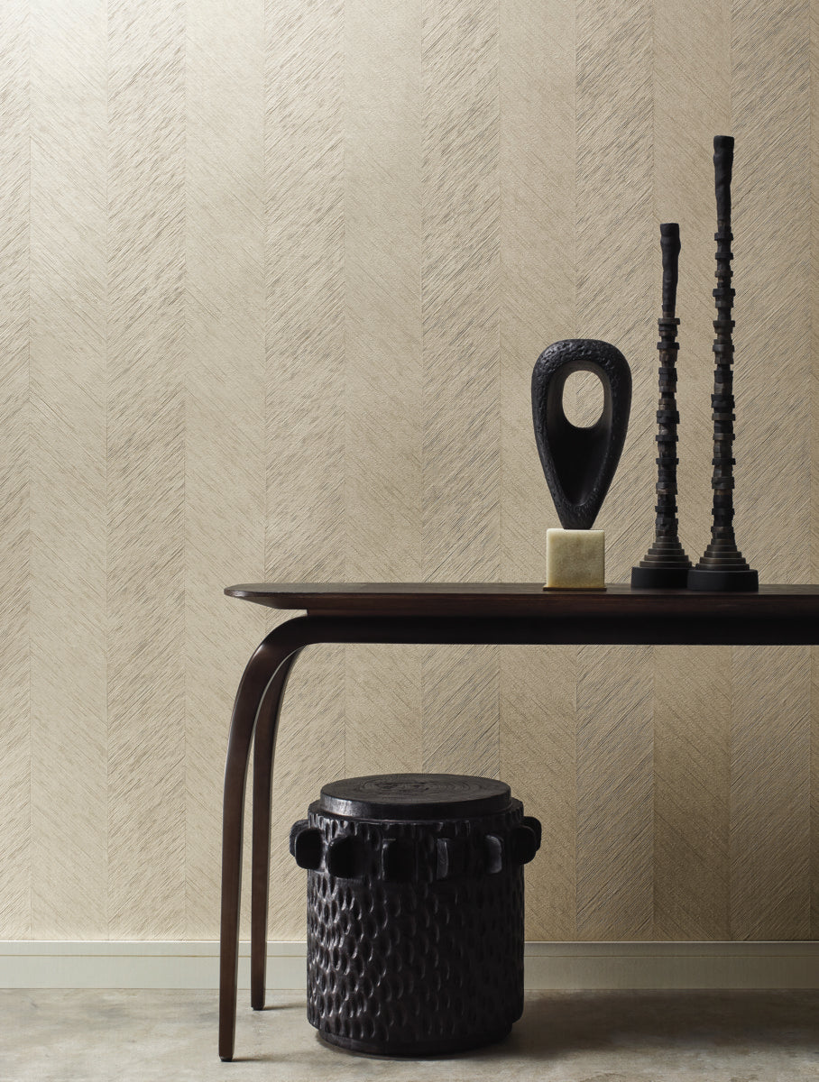 In a minimalist room, a dark wooden table is adorned with two tall black candlesticks and an abstract sculpture. Below, a textured black stool rests on a light floor against Decor2Go Wallpaper Mural's Metallic Chevron Wallpaper Cream (per Yard), highlighting its subtle pattern and tear-resistant design.