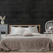 Bedroom - A minimalist bedroom features a wooden bed frame with white and beige bedding against the Midnight Slab Wallpaper Mural by Decor2Go, which provides a dark concrete-textured wall. A small blue side table adorned with flowers and a white chair paired with a basket are positioned beside the bed, adding to the urban aesthetic.