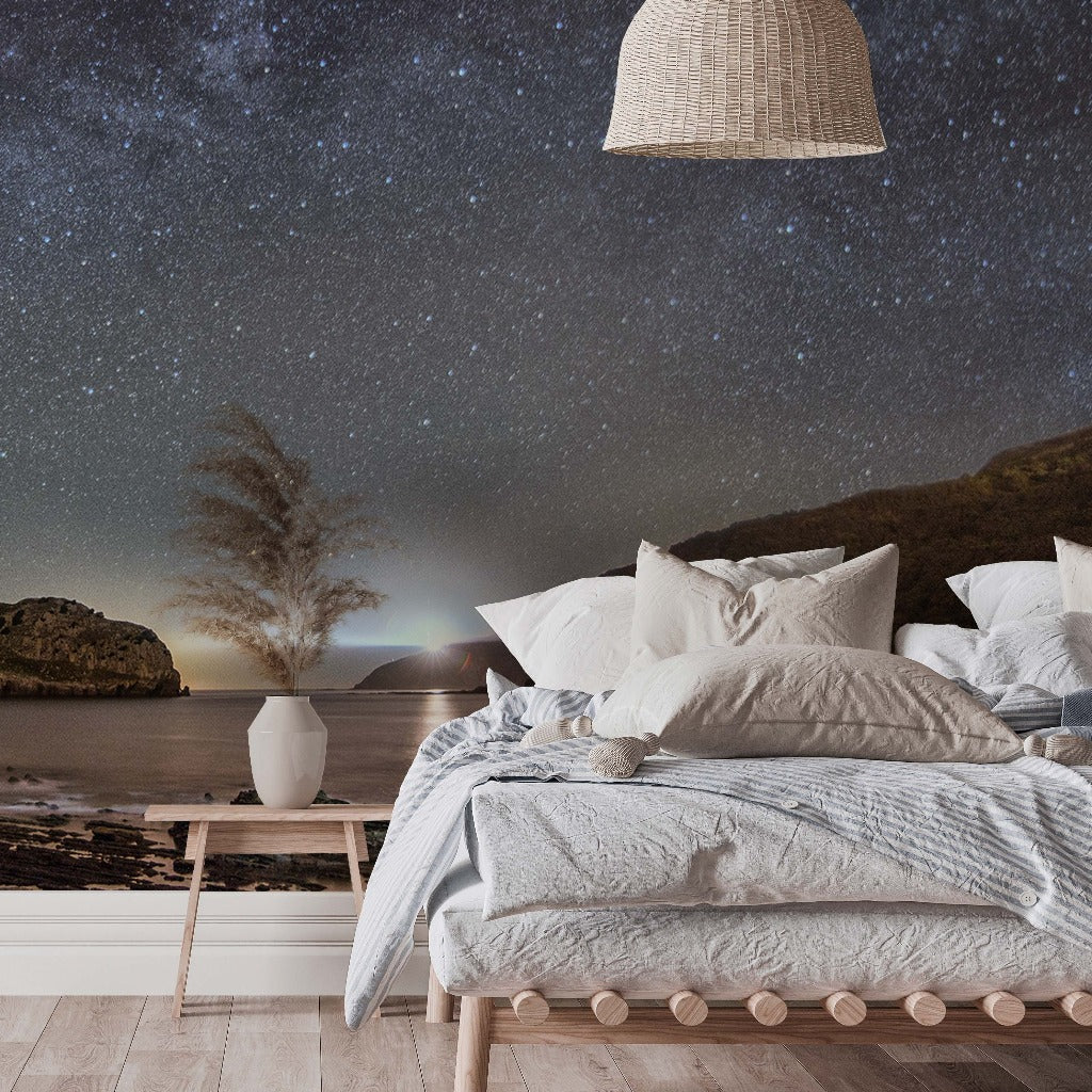 A serene bedroom with a Milky Way Galactic Coast Wallpaper Mural from Decor2Go Wallpaper Mural, featuring the Milky Way galaxy, with a cozy bed adorned with plush pillows and a gray blanket, a wicker pendant lamp above, and a side table.