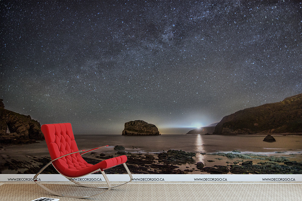 A red lounge chair on a sandy beach at night with the Milky Way Galactic Coast Wallpaper Mural from Decor2Go Wallpaper Mural highlighting the Milky Way galaxy above, and a distant lighthouse illuminating the sea and cliffs.