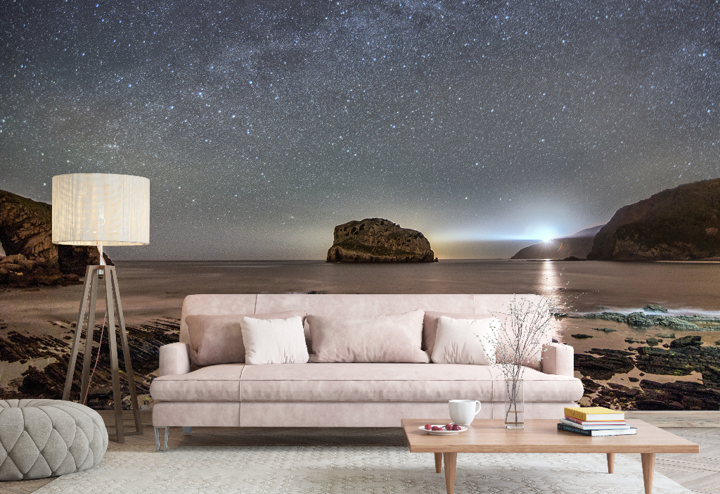 A serene living room setup with a pastel-pink couch, lamp, and astrology-themed decor items, juxtaposed against a surreal backdrop of a Milky Way Galactic Coast wallpaper mural depicting a starry sky and a moon by Decor2Go Wallpaper Mural.