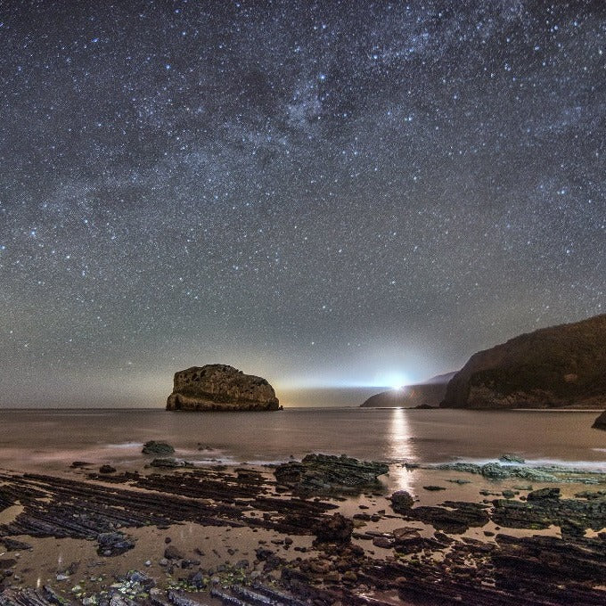 A nighttime seascape featuring a star-filled sky, showcasing the Milky Way galaxy, and a serene ocean with a large rock formation illuminated by a distant light. Rocky shores and gentle waves frame the tranquil scene, all brought to life in the Milky Way Galactic Coast Wallpaper Mural by Decor2Go Wallpaper Mural.