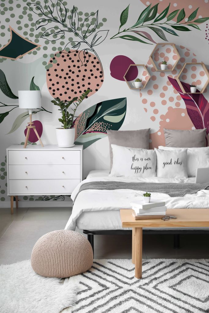 A modern bedroom with a white bed, Decor2Go Wallpaper Mural, geometric decor elements, and a cozy seating area with a pink pouf and white rug.