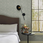 A neatly made bed with white linens sits by a round black side table with a floral arrangement, candle, and book. A modern black sconce and large window stand out against York Wallcoverings’ Mineral Fable Wallpaper (60 SqFt).