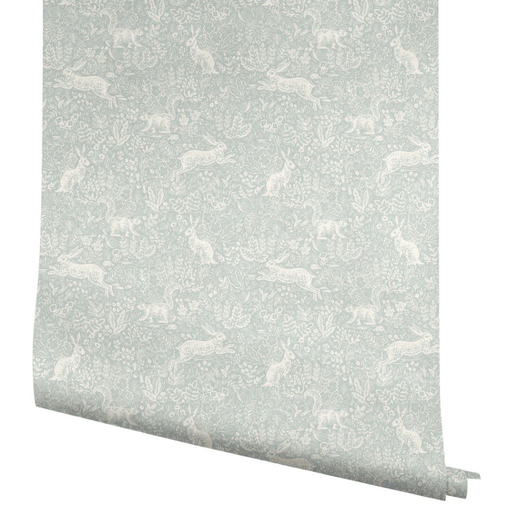 A roll of Mineral Fable Wallpaper (60 SqFt) by York Wallcoverings in light blue, featuring a subtle white pattern of rabbits, plants, and flowers in a whimsical, repeating woodland design.