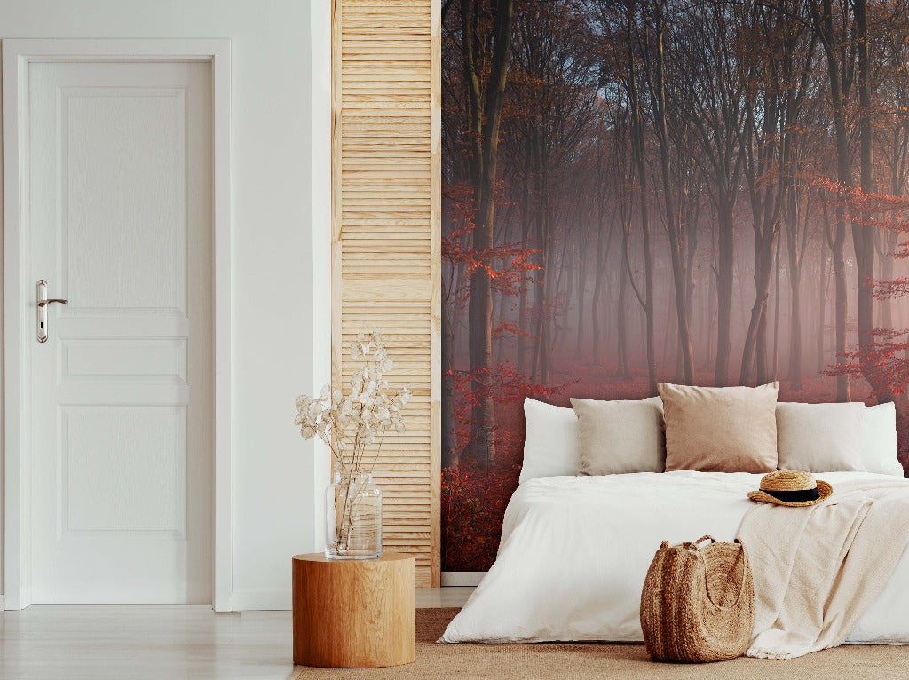 A modern bedroom featuring a white bed with beige pillows, a wooden stool with a vase and dried flowers, and a large window with a view of a Decor2Go Misty Red Forest Wallpaper Mural.
