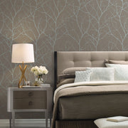 Bedroom - A stylish features a gray upholstered bed with beige bedding and decorative pillows. A modern lamp and bouquet sit on the nightstand, enhanced by York Wallcoverings' Mocha & Silver Trees Silhouette Wallpaper, showcasing an elegant light-colored tree branch pattern