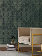 A modern living room features a beige armchair with gold accents, sitting atop a light gray rug. The elegant backdrop is provided by the dark green geometric patterned wallpaper, while a small vase adds a touch of blue to complement the space. A stack of books rests gracefully beside the chair.