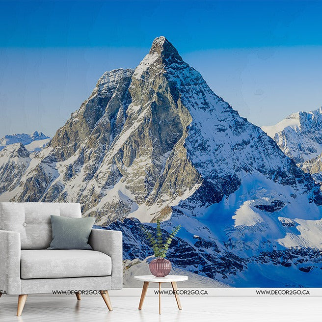 A modern gray armchair and a small plant on a wooden table set against the Decor2Go Wallpaper Mural of the majestic Matterhorn mountain range and blue sky.
