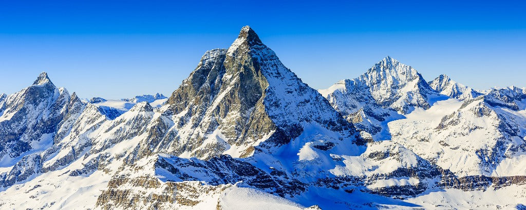 A panoramic view of the snow-covered, jagged Matterhorn mountain range under a clear blue sky, surrounded by various other sharp peaks in a rugged alpine landscape with Decor2Go Wallpaper Mural's Mountain Tops Wallpaper Mural.