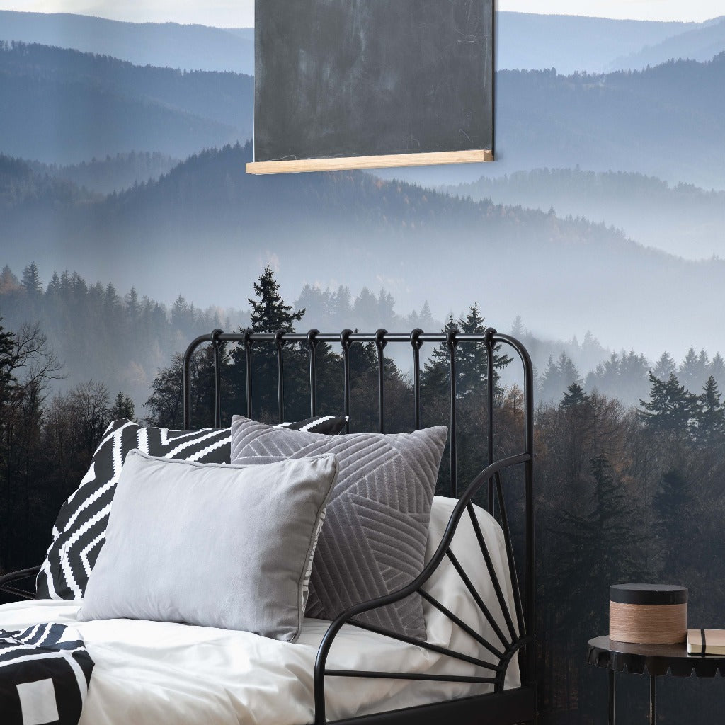 A cozy bedroom with a wrought iron bed adorned with patterned pillows, featuring a Decor2Go Wallpaper Mural, overlooking a foggy, tree-covered mountain landscape through large windows.