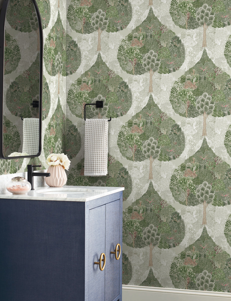 Bathroom - The includes a dark blue vanity with a white countertop, complemented by a round mirror and a textured hand towel on a black holder. The walls are adorned with York Wallcoverings' Mystic Forest Wallpaper in Green/Coral, which features green leaf and tree patterns that enhance the nature-inspired decor. A small vase with flowers is elegantly placed on the counter