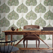 Home Office - A cozy space includes a wooden desk and a chair with a leather backrest, adorned with an open book, decorative pieces, and a geometric lamp. The room is decorated with the Mystic Forest Wallpaper by York Wallcoverings in green and coral tones