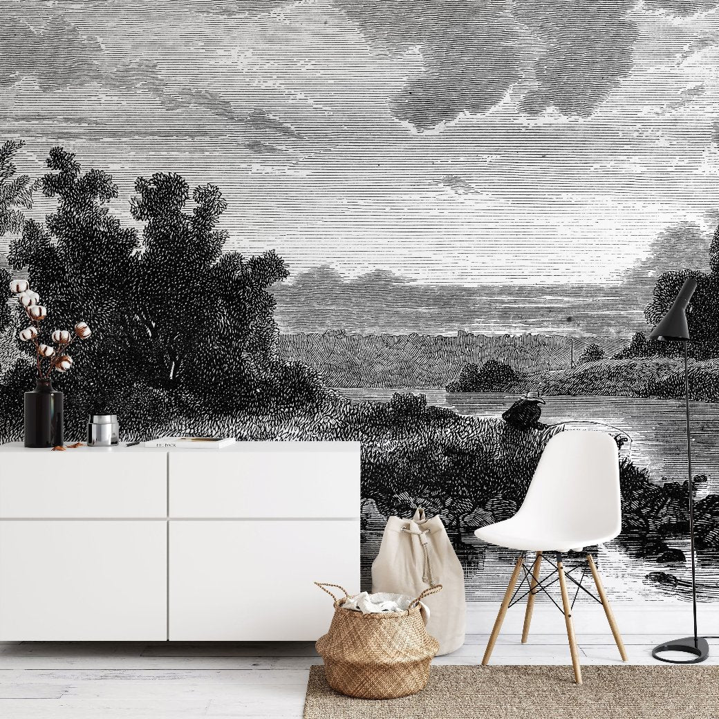 A minimalist room showcases Decor2Go Wallpaper Mural's Mystic Lake Wallpaper Mural, featuring a black and white vintage landscape with trees and a river. The scene complements a white chair, black floor lamp, white cabinet, and woven basket beside cotton stems in a vase.