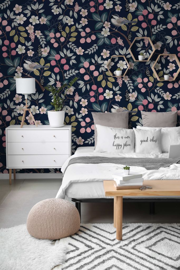 Bedroom - A cozy bedroom featuring a feature wall with Decor2Go Wallpaper Mural, white bedding with decorative pillows, a white and wood nightstand, a small lamp, and a textured rug. A patterned ottoman sits in