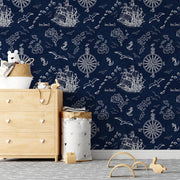 Kids Room - A children's room featuring a wooden dresser and toy storage is adorned with the Nautical Treasure Adventure Wallpaper Mural by Decor2Go, showcasing ships, compasses, and birds on a navy background. A small wooden arch toy rests on the floor beside a cozy rug, inviting little ones to dream of pirate adventures.