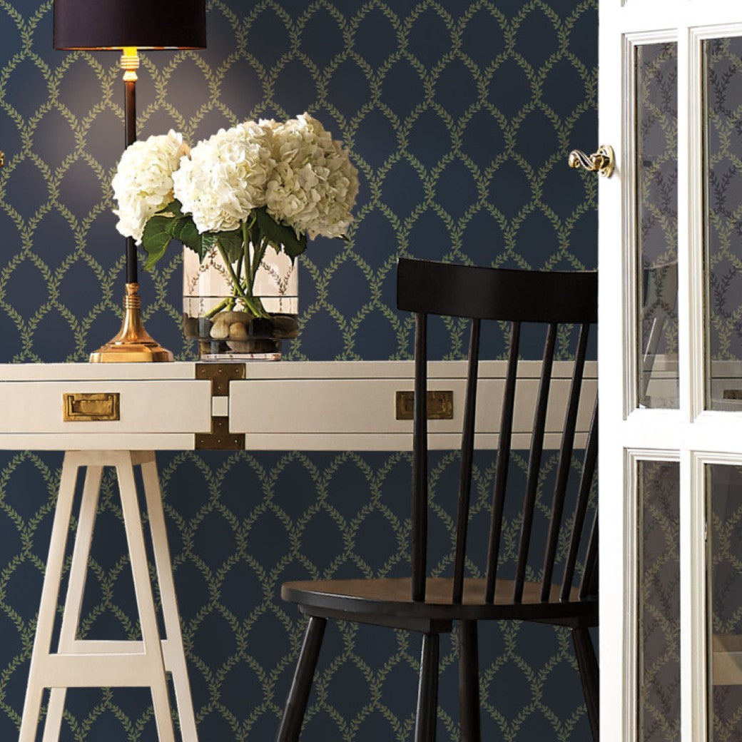 A white desk with drawers and a black chair sit in front of York Wallcoverings’ Navy Laurel Wallpaper. The desk holds a lamp with a black shade and a vase of white hydrangeas. A glass door nearby is partially open.