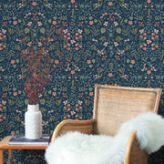 A rattan chair with a white fur throw sits by a table topped with a white vase of dried branches, set against York Wallcoverings' Navy Wildwood Wallpaper (60 SqFt).