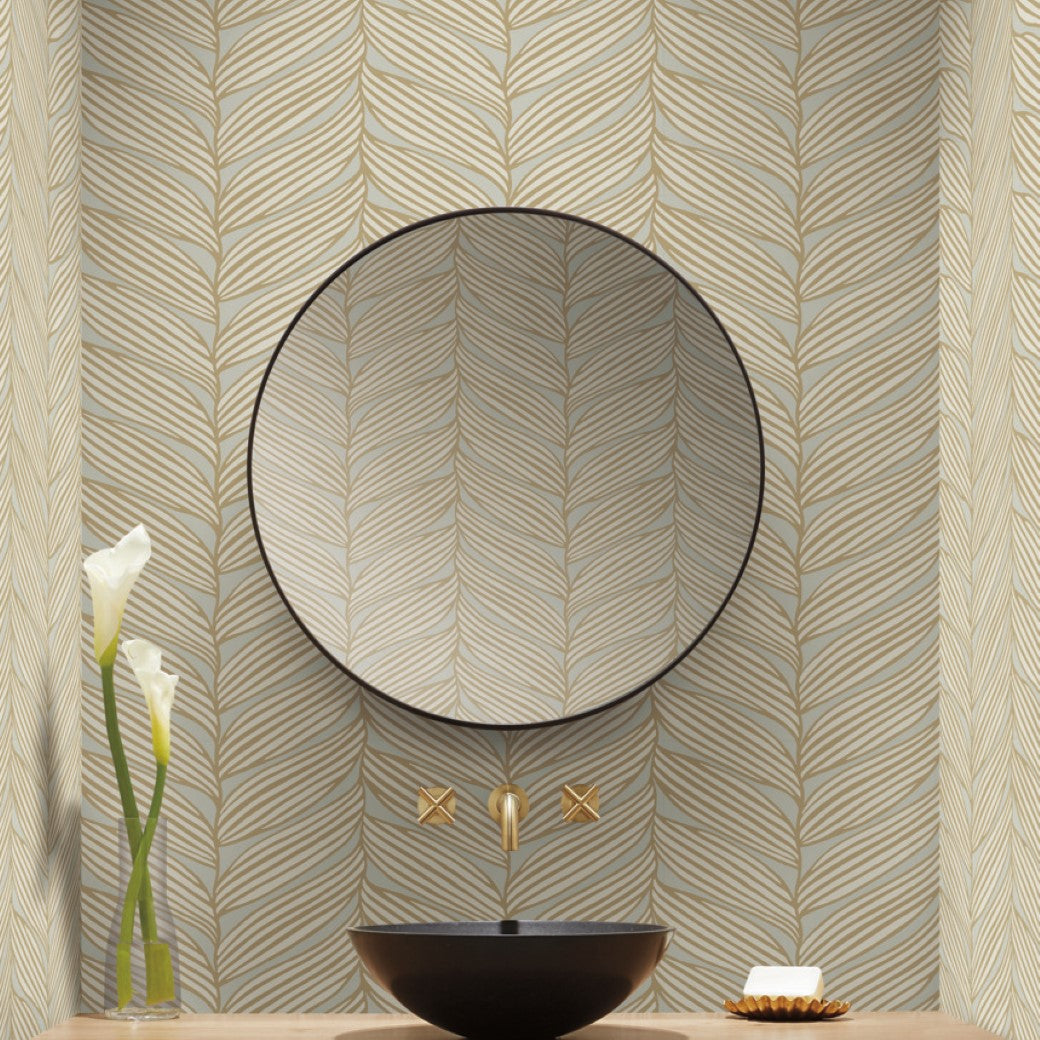 Bathroom - A has a round mirror above a black vessel sink with a gold faucet from the Modern Metals Collection, set against York Wallcoverings' Neutral & Gold Luminous Leaves Wallpaper. Two white calla lilies in a tall vase to the left add elegance