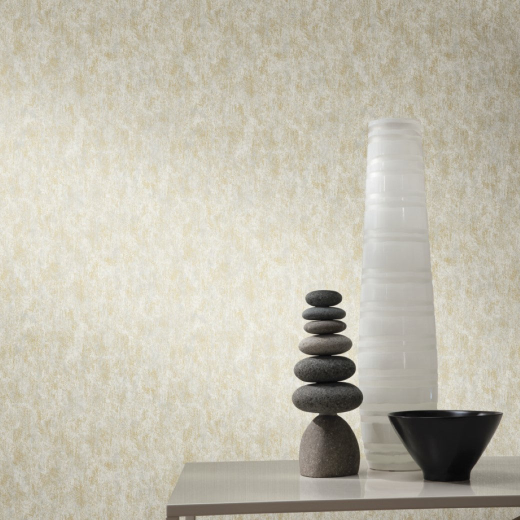 A minimalist design with stacked stones, a tall vase, and a black bowl on a light tabletop. The space is enhanced by contemporary interiors featuring York Wallcoverings' Neutral & Gold Shimmering Patina Wallpaper against a textured beige wall.