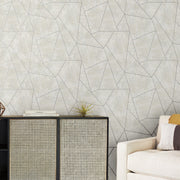 Living Room - A modern features York Wallcoverings' Neutrals & Gold Metallic Cascade Wallpaper with a geometric pattern, a cabinet with cane doors and a plant on top, and a cream sofa partially visible with a brown and white pillow