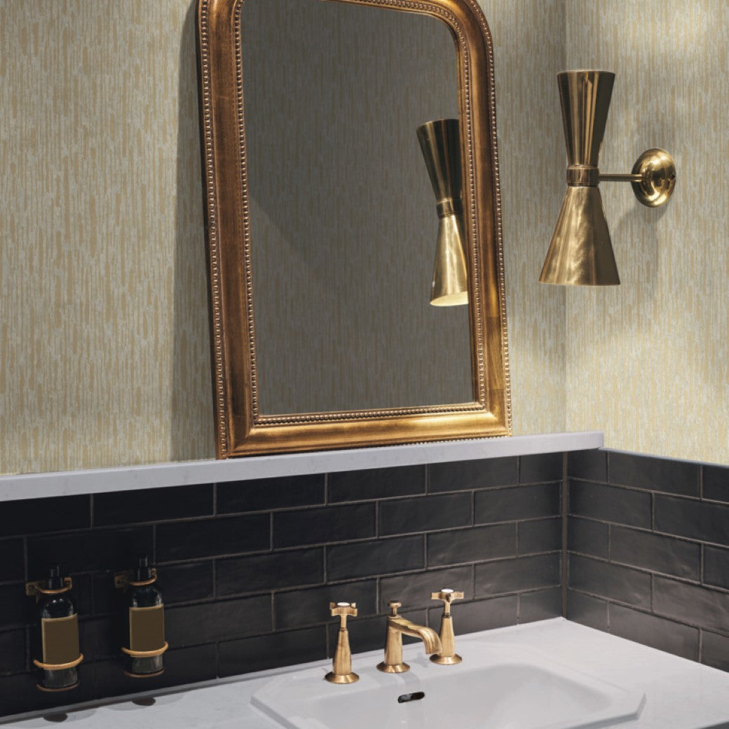 Bathroom - A showcases a large gold-framed mirror above a sink with matching gold faucet and handles. The brass wall light and dark tiles contrast the York Wallcoverings Neutral & Silver Nazca Wallpaper, enhancing the modern look that borders the white countertop